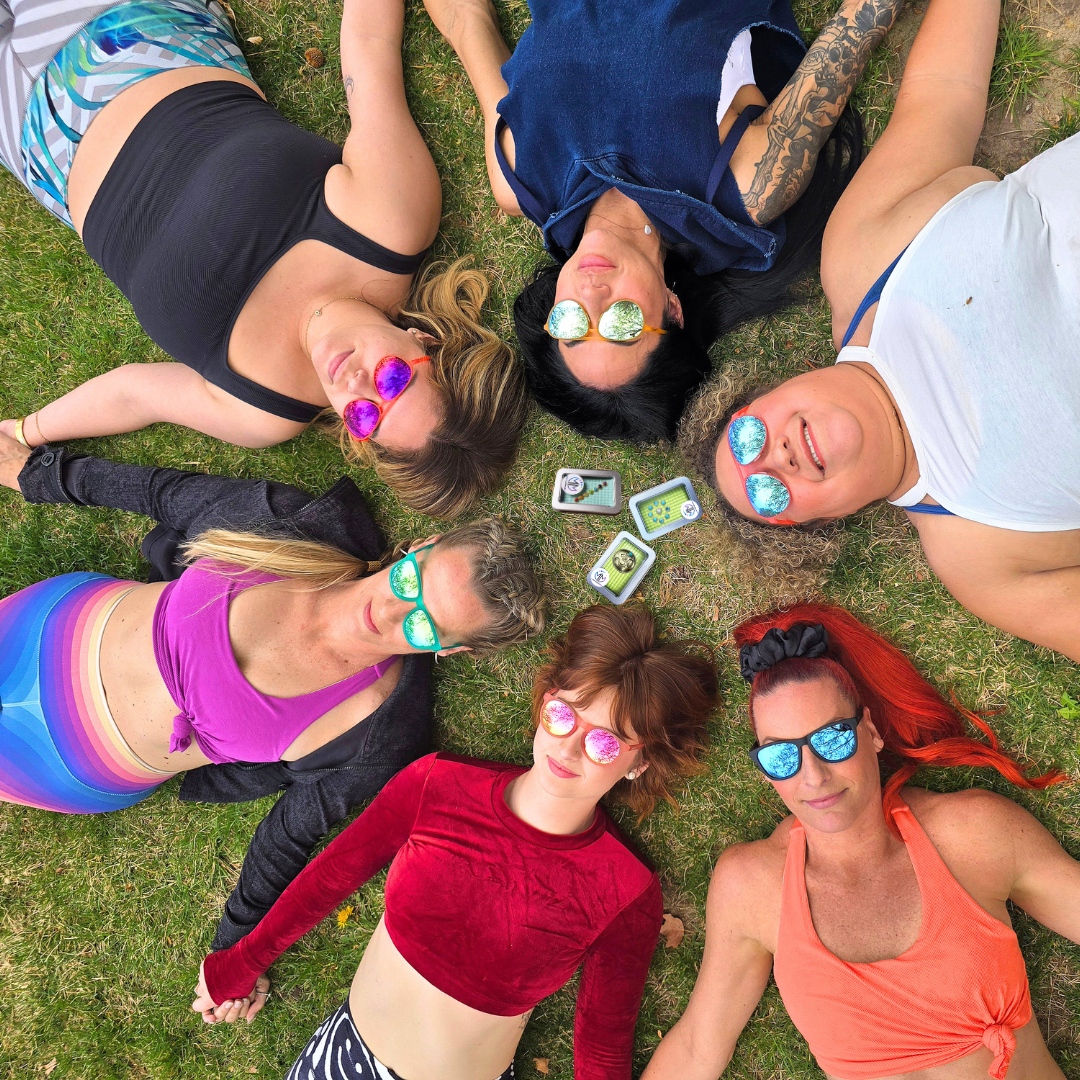 5 woman laying in the grass holding hands with yoga mat charms in the center. They are all wearing sunglasses and yoga outfits