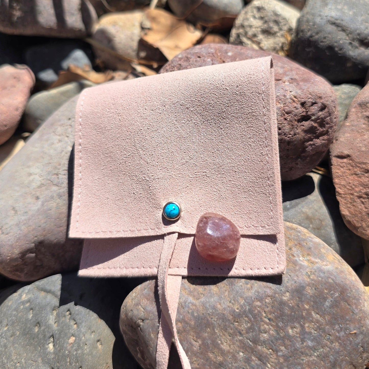 pouch to hold crystals in with a turquoise stone on the front. there is a a stone sitting on the bag and the back ground is river rocks