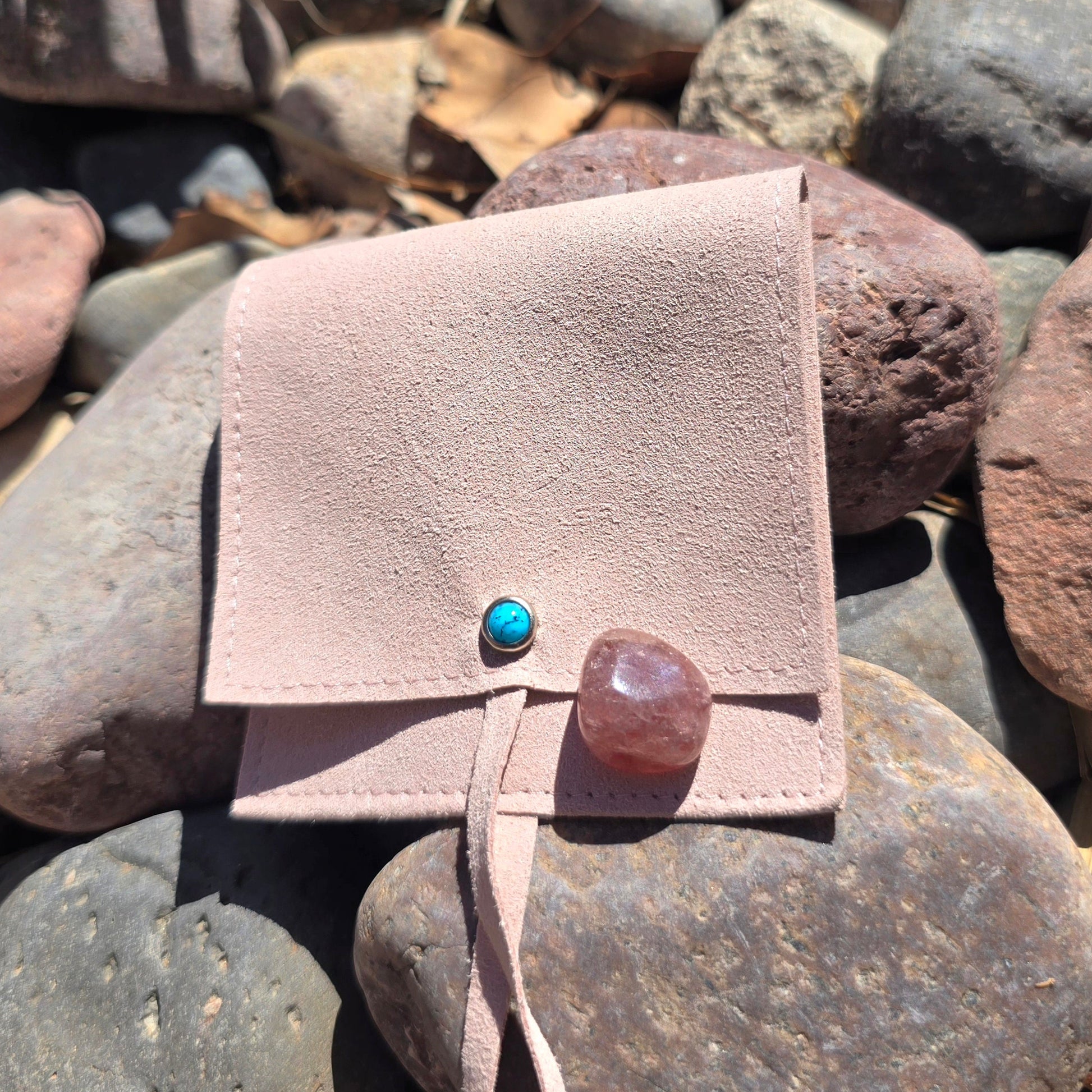 pouch to hold crystals in with a turquoise stone on the front. there is a a stone sitting on the bag and the back ground is river rocks